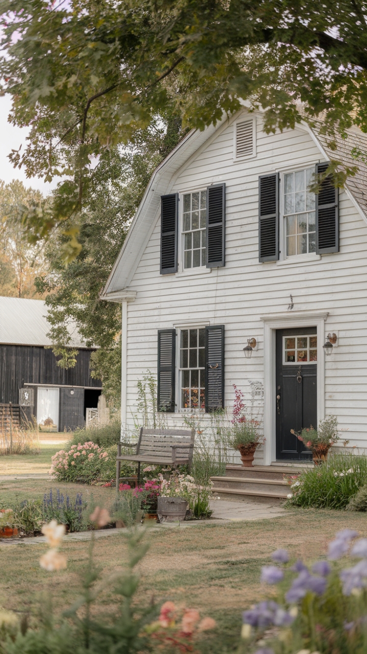 A charming vintage farmhouse exudes timeless appeal with its crisp white exterior contrasted by bold black accents on the door and shutters. The inviting yard blooms with a vibrant tapestry of flowers, adding a touch of natural elegance. A rustic wooden bench beckons for quiet moments, while the backdrop of a classic barn and lush trees completes this idyllic country retreat. The overall scene captures a perfect blend of rustic charm and farmhouse chic, inviting warmth and nostalgic sophistication.